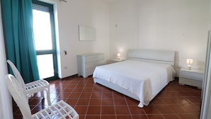 A bedroom with a bed, nightstands, a chair, a window with curtains, and a cross on the wall.
