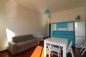 Un espace de vie compact comprenant un canapé, une table à manger, des chaises, un petit coin cuisine avec un réfrigérateur et des placards, ainsi qu’un ventilateur de plafond.