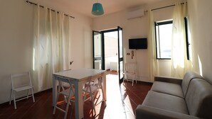 A room with a table and chairs, a sofa, a TV mounted on the wall, and a window with curtains.