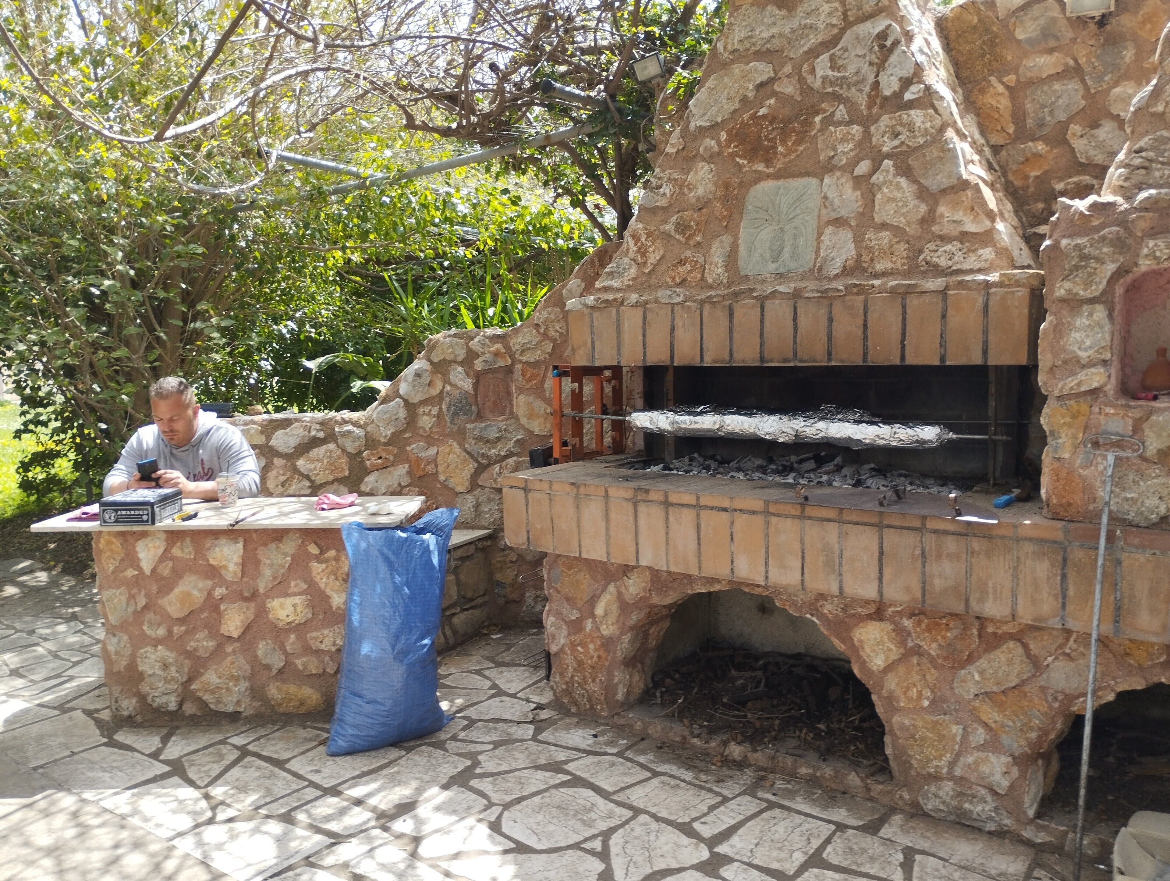 A stone-built outdoor oven with a brick base, a person sitting at a table with a laptop, and a blue sack on the ground.