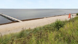Een strand met een betonnen pad dat naar het water leidt, een zandstrand en wat begroeiing op de voorgrond.