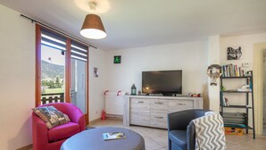 Una sala de estar con televisor, dos sillones, un sofá, una mesa, una estantería y vistas al exterior.