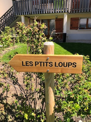 Un cartel de madera con la inscripción 'LES PTITS LOUPS', sobre un fondo de jardín con un edificio al fondo.