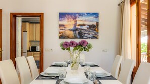 Un comedor con una mesa dispuesta para cuatro, un florero con flores y un cuadro de una puesta de sol sobre rocas.