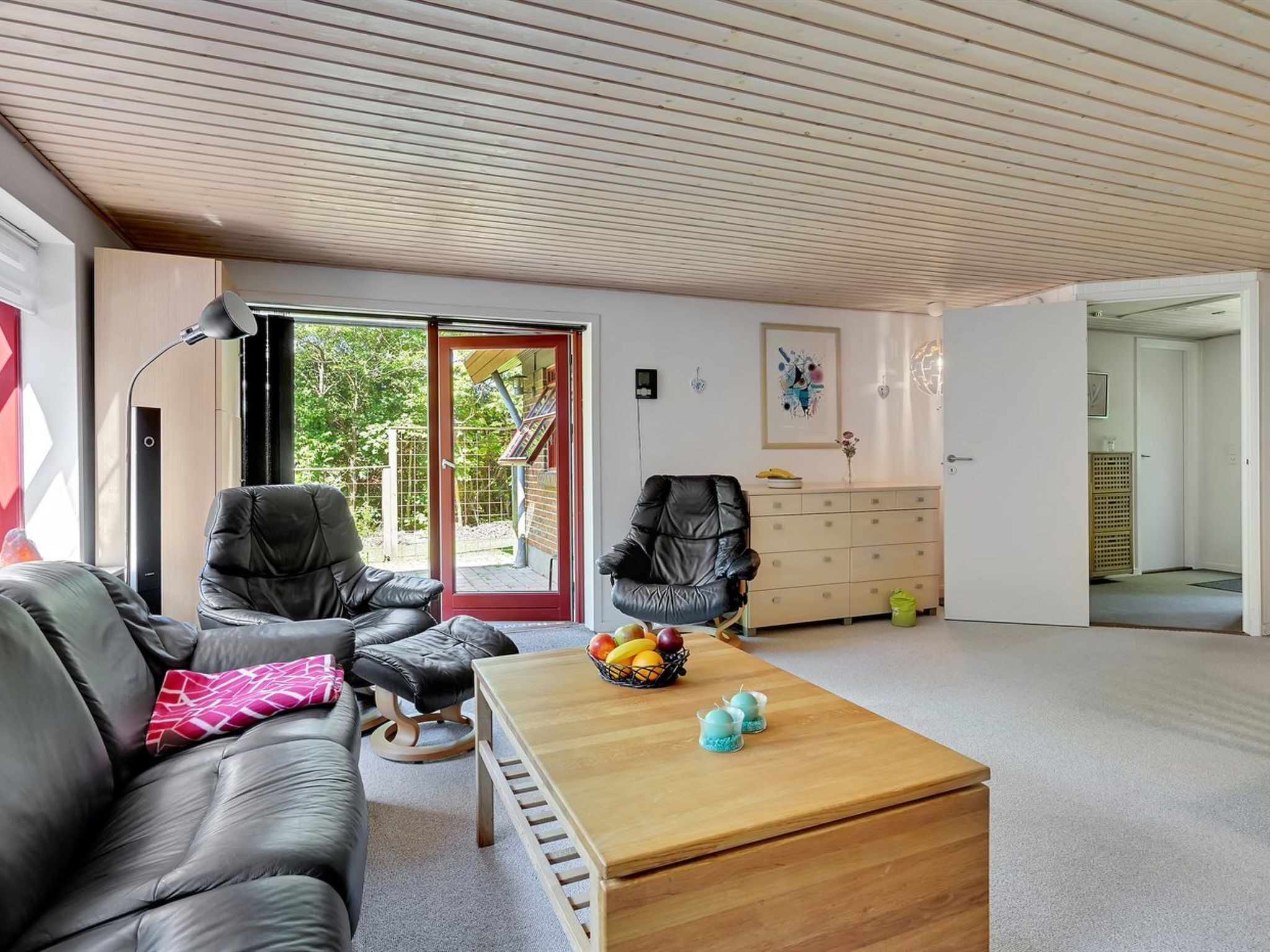 A modern living room with a leather sofa, a wooden coffee table, and a black leather armchair. There is a red door leading to an outdoor area, and a white dresser with drawers.