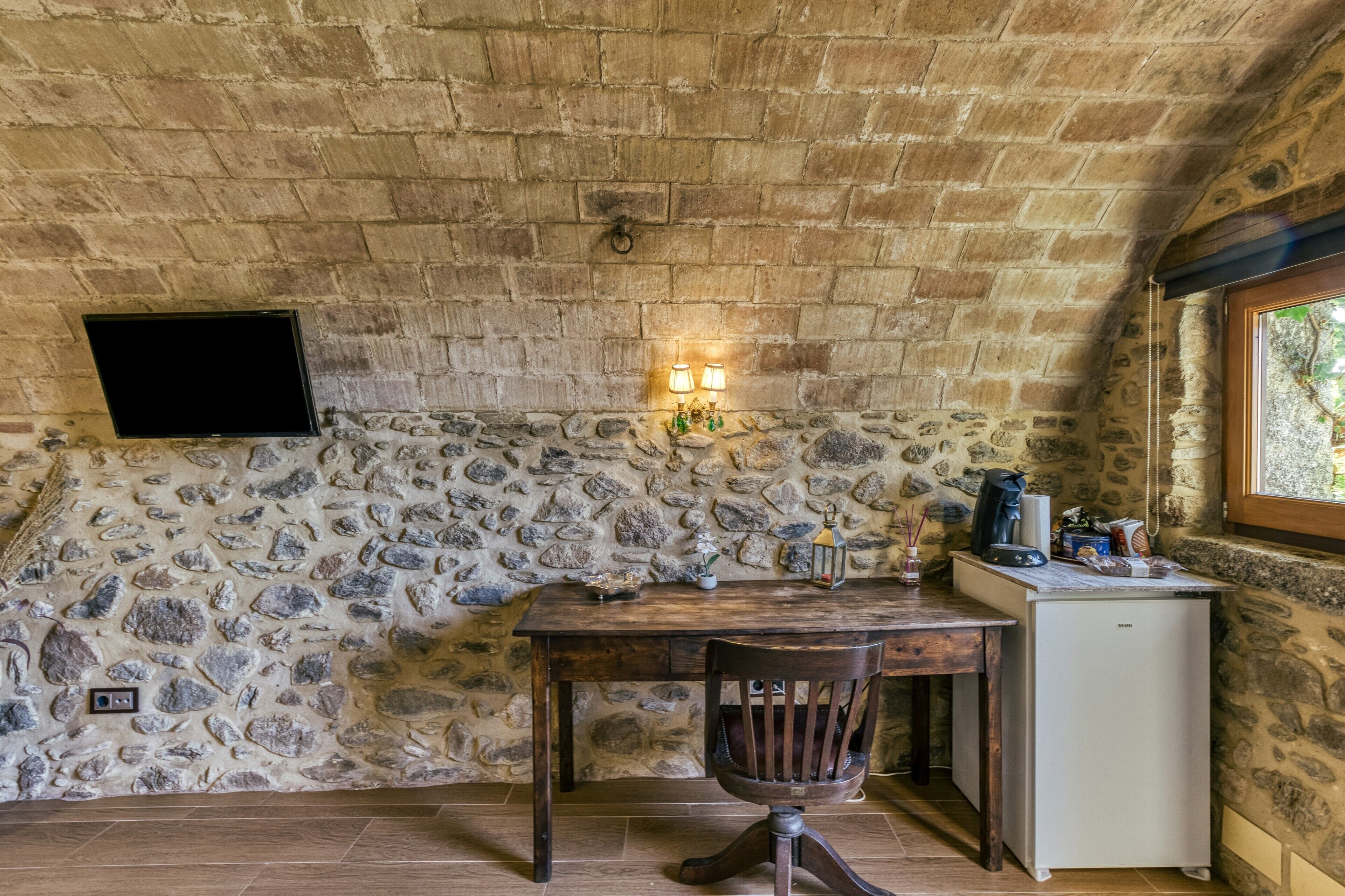A rustic room with a stone wall, a wooden table, a chair, a small white refrigerator, and a TV mounted on the wall.