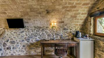 A rustic room with a stone wall, a wooden table, a chair, a small white refrigerator, and a TV mounted on the wall.