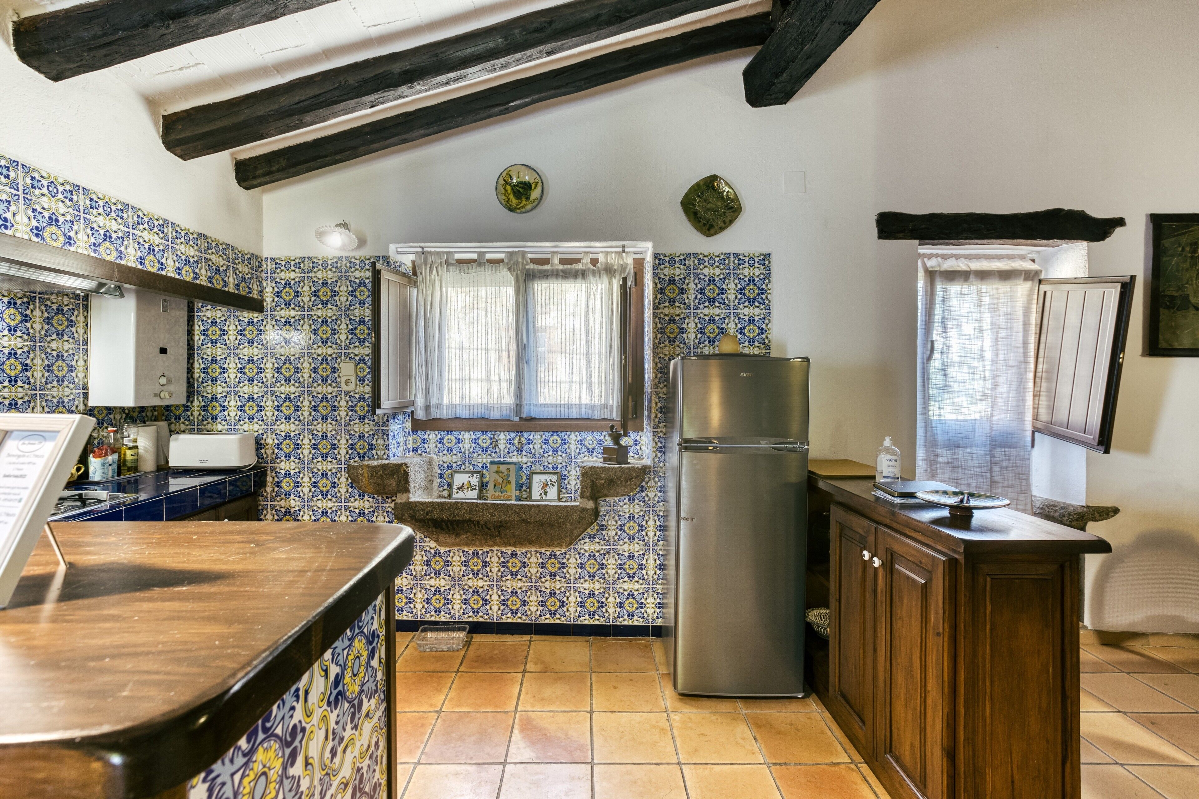 Een keuken met blauwe en gele patroontegels, een roestvrijstalen koelkast, houten kasten en een raam met dunne gordijnen.