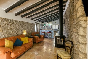 A rustic interior with exposed beams, a stone wall, a tiled floor, a sofa with cushions, a wooden chair, and a kitchen area with a stove. - Vacation Home "Can Cortada" with Mountain View, Private Terrace & Barbecue (Cantallops)