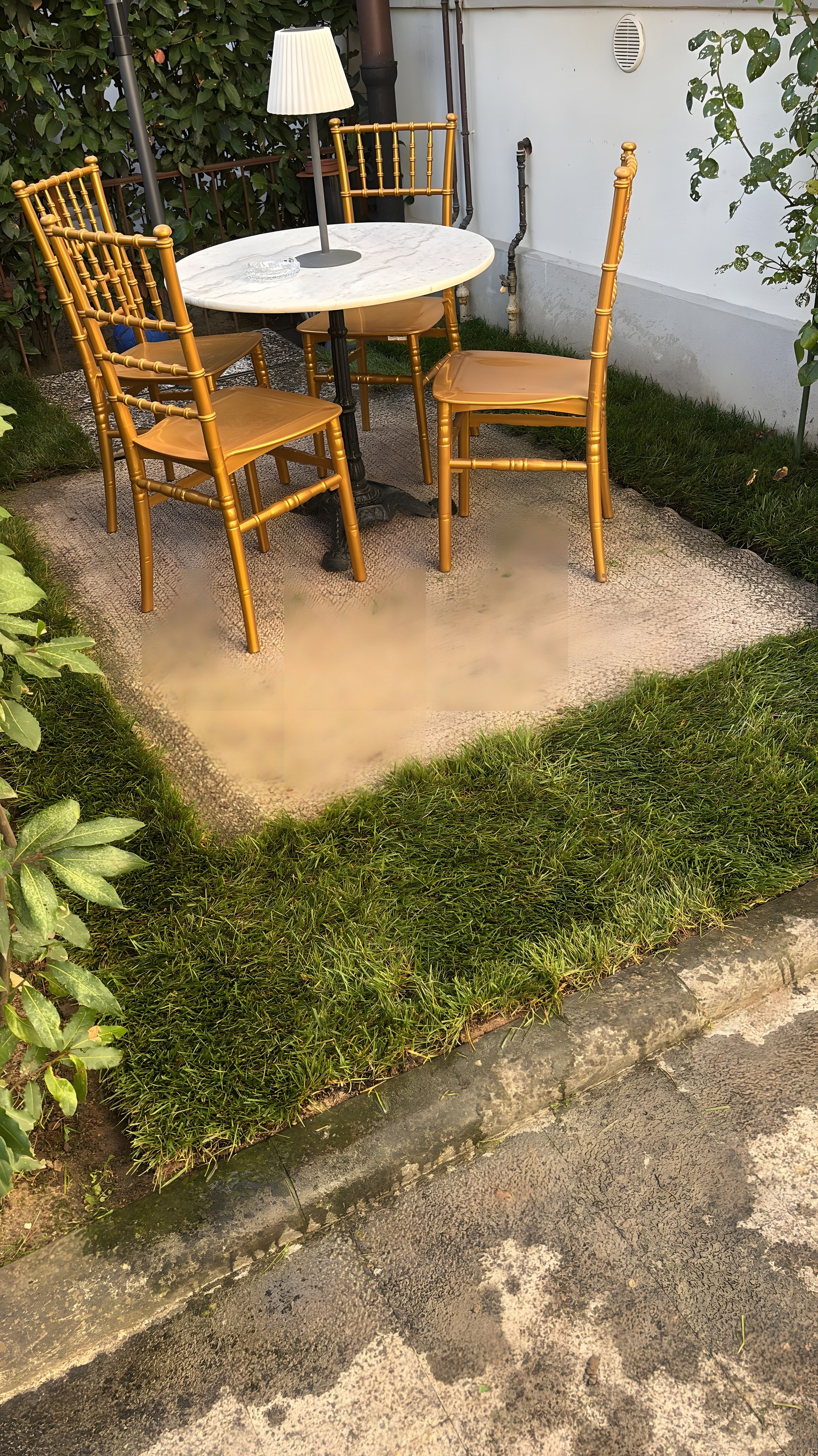 Un'area salotto all'aperto con un tavolo rotondo e quattro sedie, circondata da un vialetto sterrato e vegetazione.