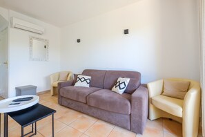 Een woonkamer met een bruine bank, twee beige fauteuils, een kleine tafel met afstandsbedieningen, een grijze poef, een witte airconditioner en een spiegel aan de muur.