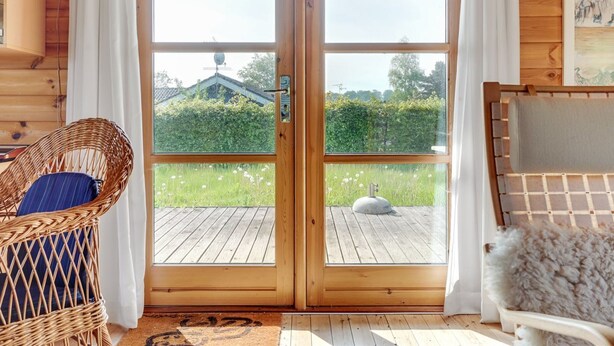 Une cabane en bois dotée d’une grande porte vitrée donnant sur une terrasse. À l’intérieur, on trouve un fauteuil en osier et une chaise blanche avec un coussin moelleux.