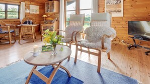 L’intérieur d’un chalet accueillant, avec des murs et des planchers en bois, une table ronde en bois ornée d’un vase de fleurs, deux chaises en osier, un téléviseur et une grande fenêtre avec des rideaux.