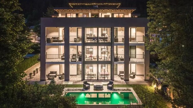 A modern multi-story building with a swimming pool, outdoor seating, and a well-lit interior.