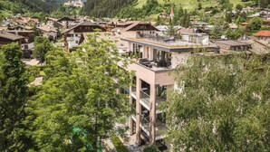 A modern building with balconies, surrounded by traditional houses and a mountainous backdrop.