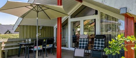 An outdoor patio with a wooden deck, a covered seating area with a table and chairs, and a large umbrella providing shade.