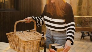 A person at an outdoor wooden table with a wicker basket, a bottle, and food items.