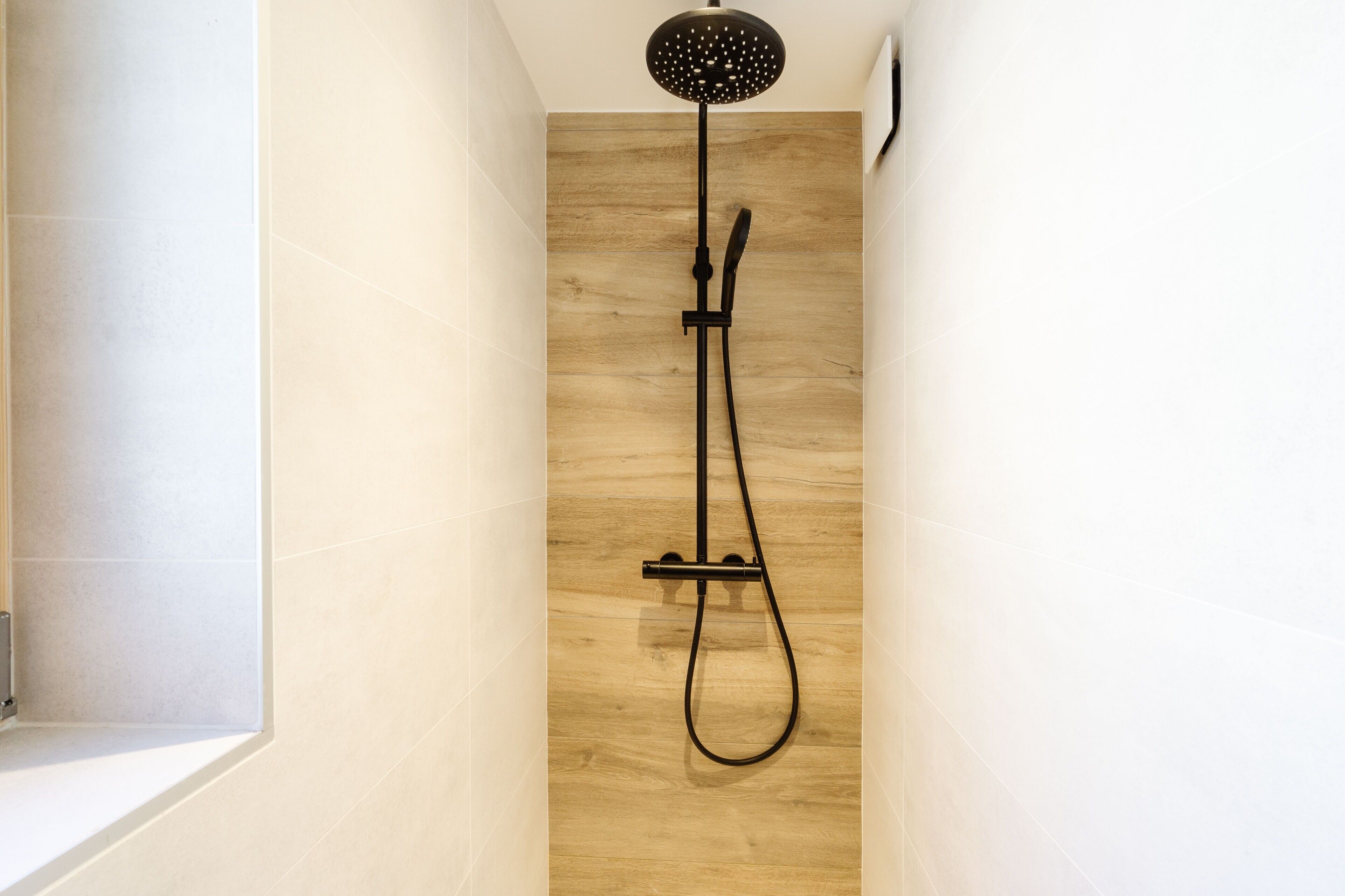 A shower with a handheld showerhead and a handheld shower attachment in a narrow, tiled room.
