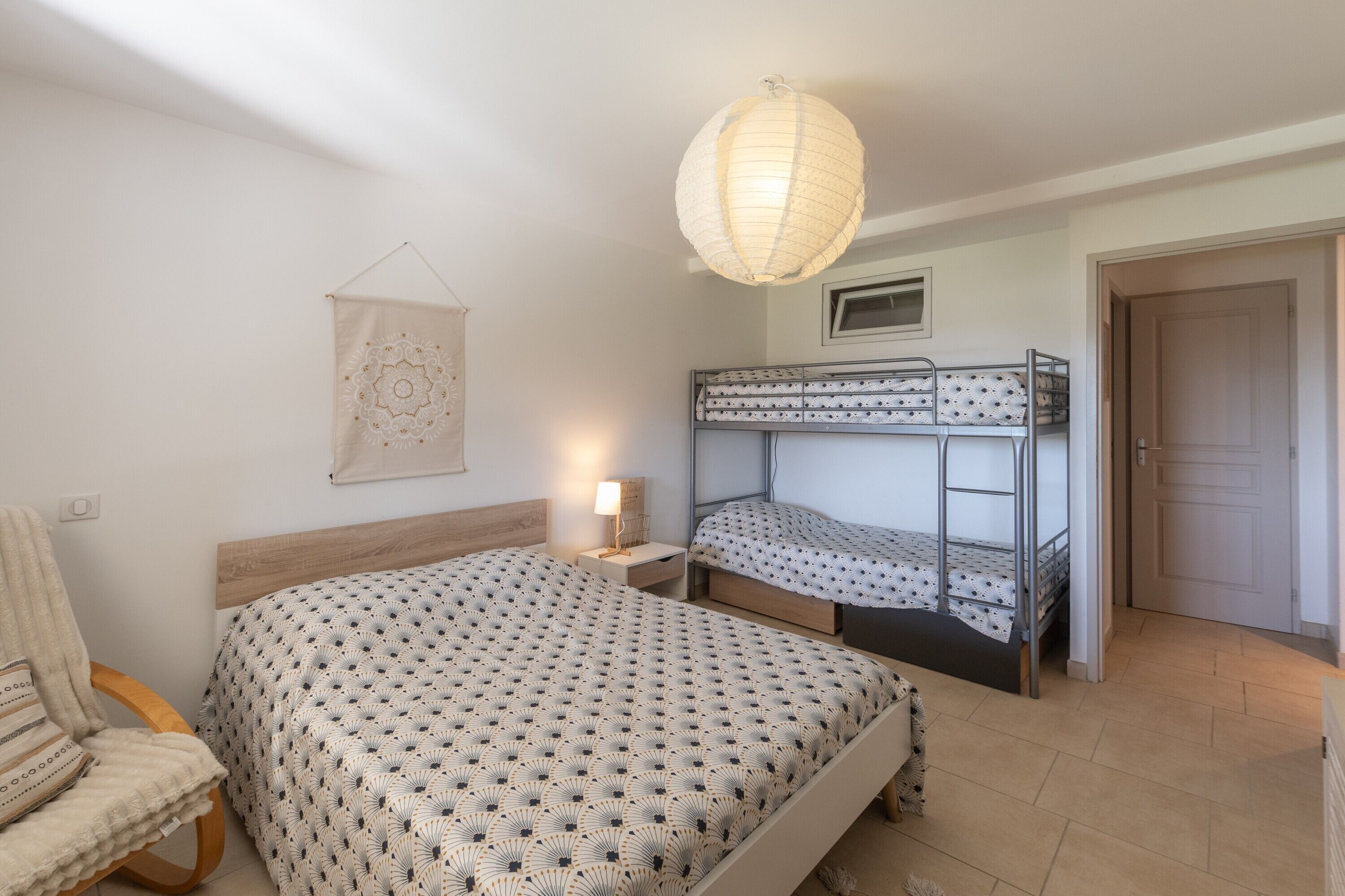 Une chambre avec des lits superposés, une chaise, une table de chevet avec une lampe, une décoration murale et une porte.