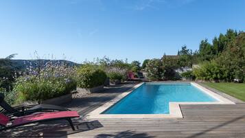 A rectangular swimming pool with a wooden deck, surrounded by lush greenery and a clear blue sky.