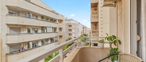 Un balcón con una silla de mimbre y una pequeña mesa, con vistas a un edificio residencial de varios pisos con balcones y macetas.
