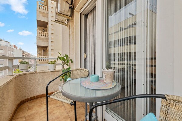 En balkon med et rundt glasbord, fletstole, krukker med planter og udsigt til omkringliggende bygninger.