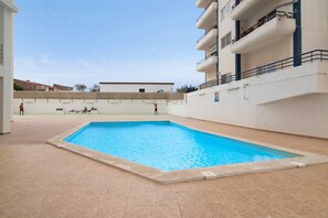 Área de piscina ao ar livre com piso de azulejos, um edifício com varandas e céu claro.