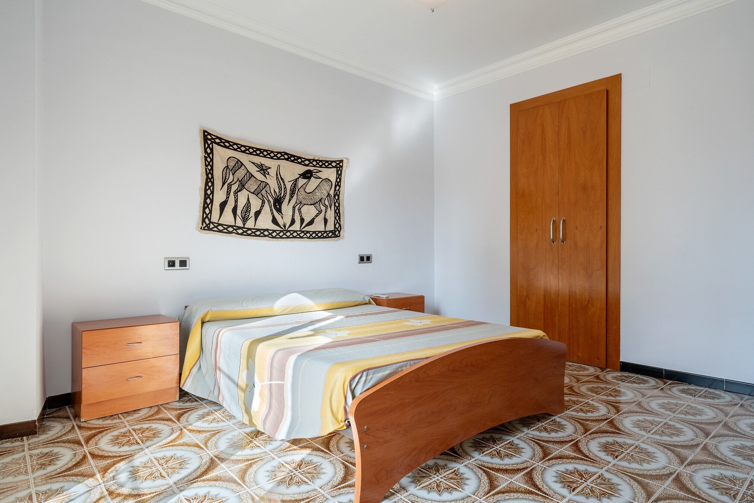 Un dormitorio con una cama, un armario de madera, una mesita de noche, un tapiz colgado en la pared y baldosas de suelo estampadas.