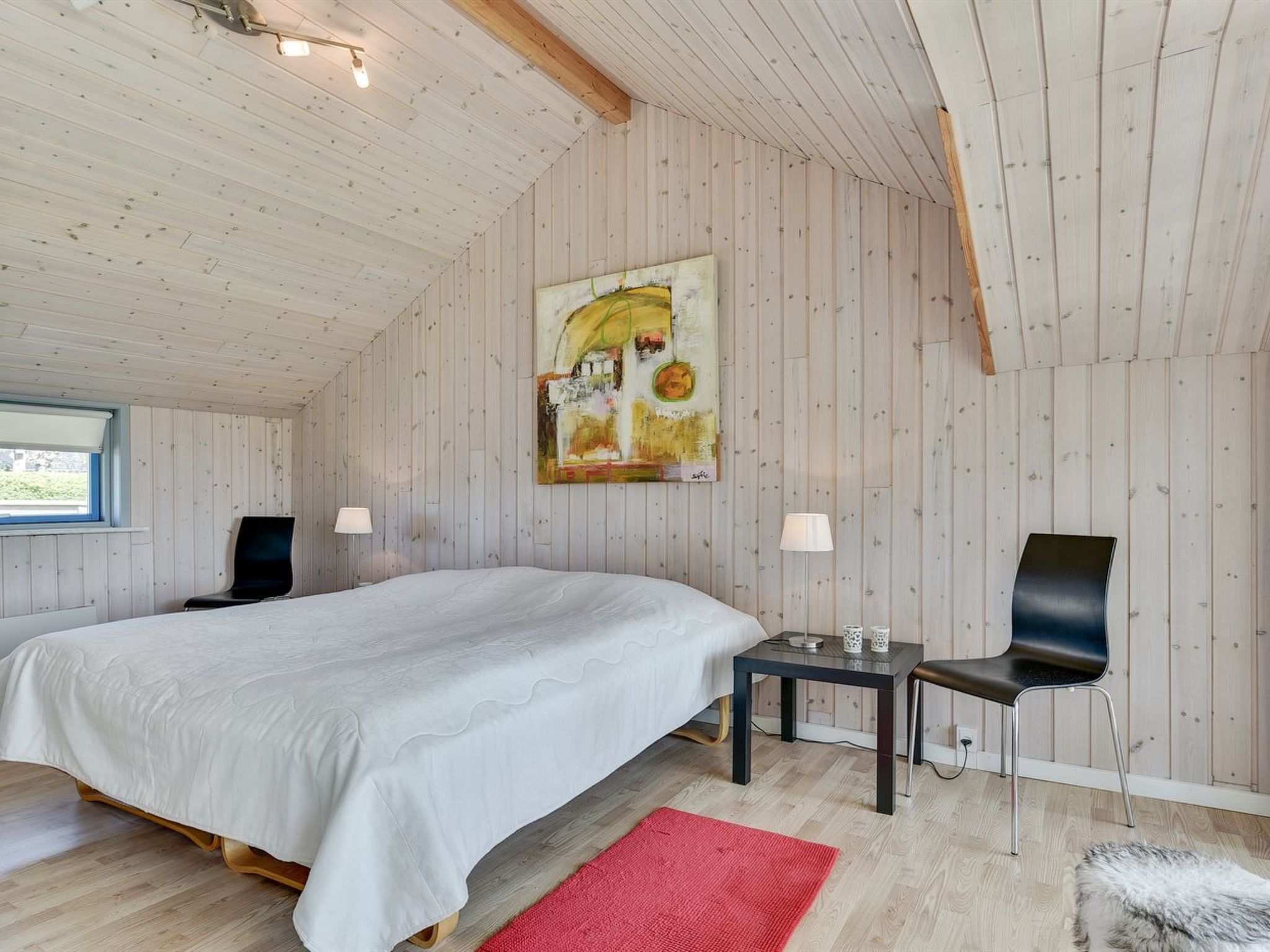 Un dormitorio con una cama, dos sillas, una mesita, un tapete rojo y un cuadro en la pared.
