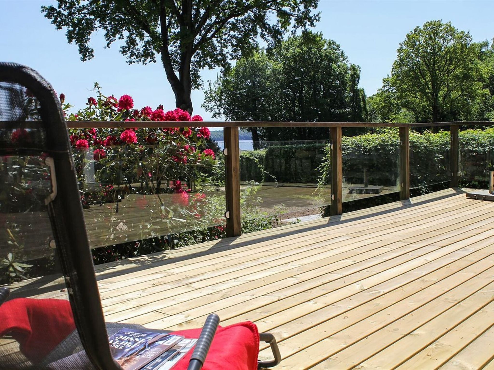 A wooden deck with a glass railing, surrounded by lush greenery and flowering plants.