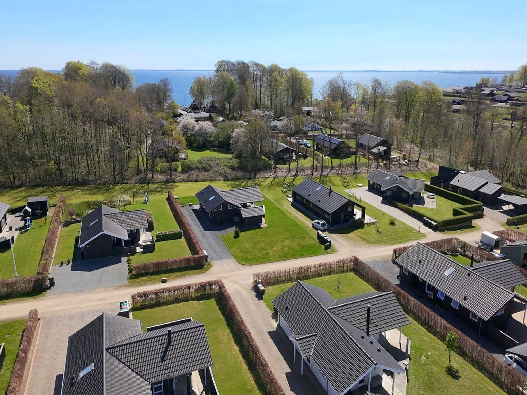 A residential area with houses arranged in a grid pattern, featuring a mix of single and double-story buildings, surrounded by greenery and a body of water in the background.