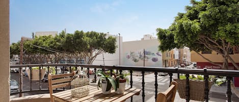 A balcony with a wooden table and chairs, potted plants, and a view of the street and buildings.