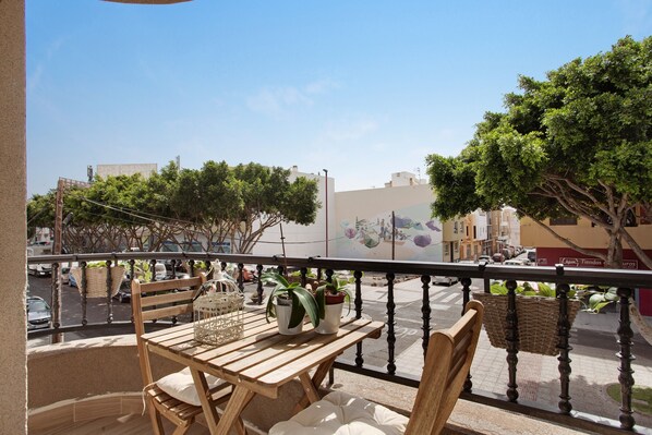 A balcony with a wooden table and chairs, potted plants, and a view of the street and buildings.