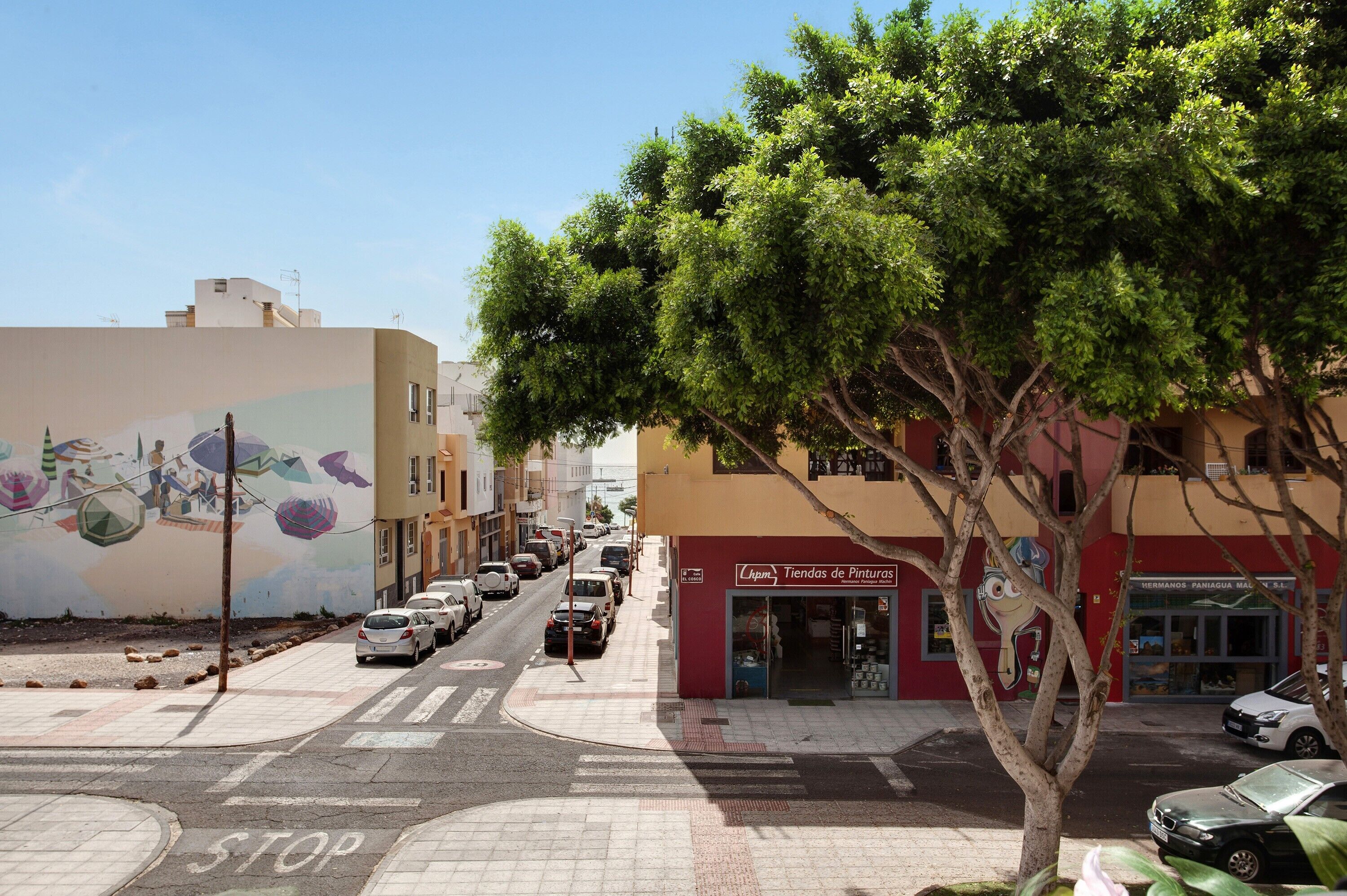 Calle con un mural, coches estacionados y una tienda llamada 'Tiendas de Pinturas'.