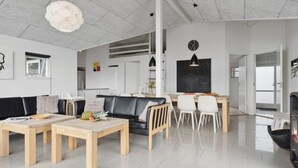 A modern living room with a black leather sofa, wooden coffee table, and white chairs. There is a dining area with a blackboard and a clock on the wall.