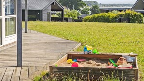 A wooden deck leading to a modern building with a glass facade, a grassy area with a sandbox, and a clear view of the surrounding landscape.