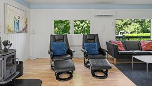 Une pièce comprenant deux fauteuils inclinables, un canapé, une table, une cheminée et de grandes fenêtres donnant sur un paysage verdoyant.