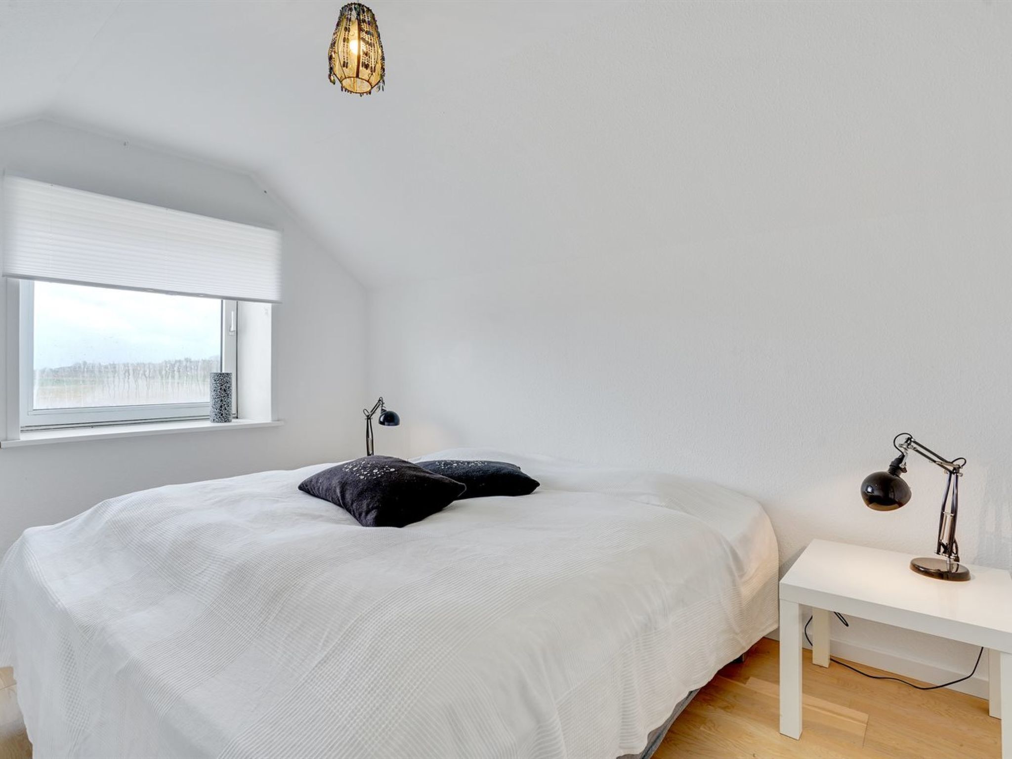 A bedroom with a bed, a bedside table, a lamp, a window with blinds, and a hanging light fixture.