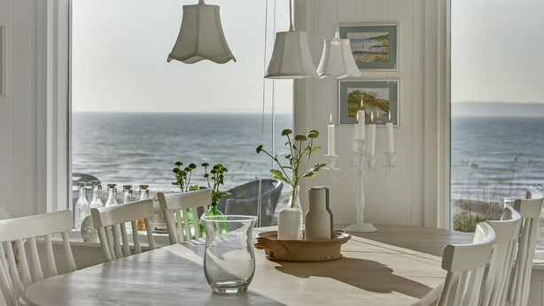 Comedor con vista al mar, muebles blancos, lámparas colgantes y una mesa con un florero y velas.