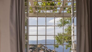 Une chambre dotée d’une grande fenêtre donnant sur une ville côtière, d’un lit en bois et offrant une vue sur la mer et les collines.