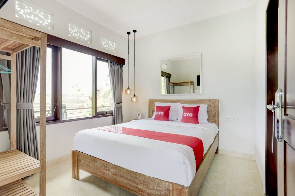 A bedroom with a wooden bed, a large window with curtains, a wooden bench, and a hanging light fixture.