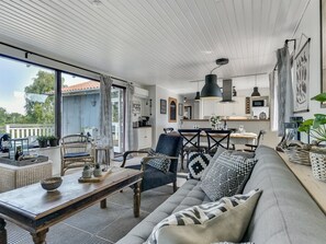 Ein großzügiger Wohnbereich mit einer Couch, Sesseln, einem Couchtisch und einem Essbereich mit Tisch und Stühlen. Ein großes Fenster sorgt für natürliches Licht und bietet einen Ausblick ins Freie.