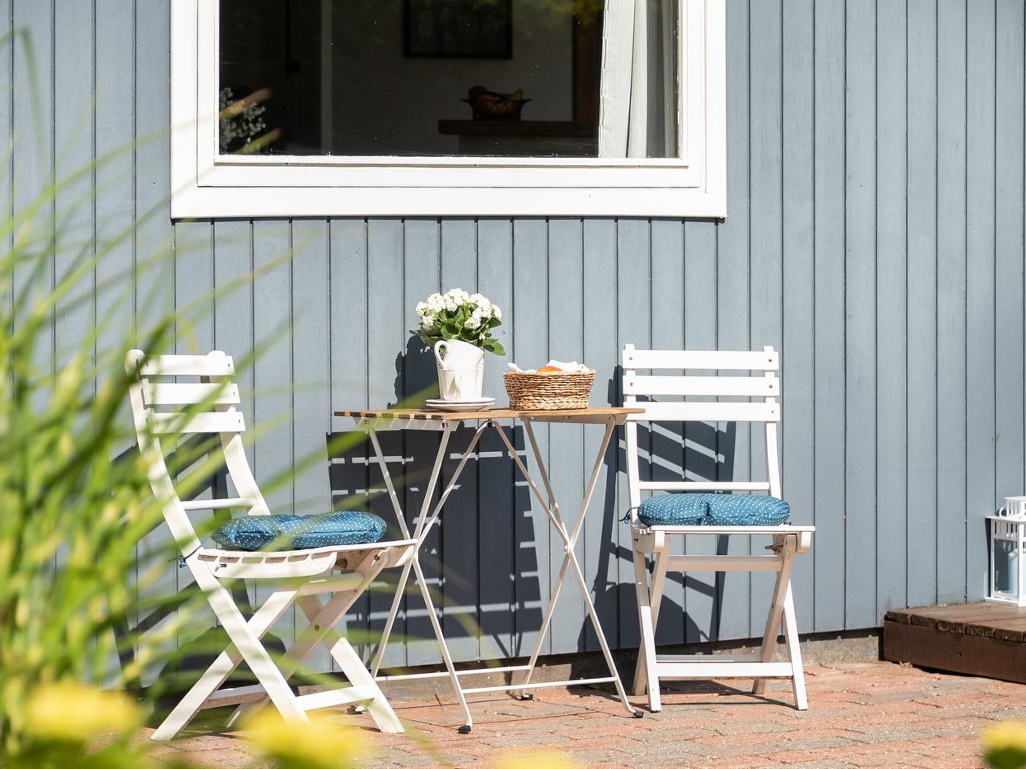 A patio with two white chairs and a table, a wicker basket, and a white pitcher with flowers.