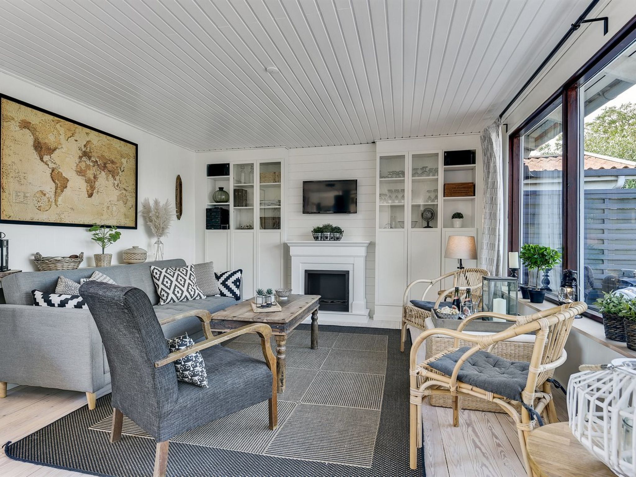 Un salon spacieux avec un canapé gris, des fauteuils en osier, une cheminée et une grande carte du monde accrochée au mur.
