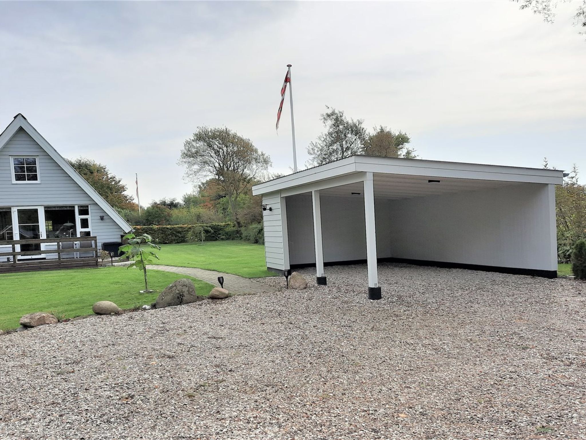 A covered outdoor area with a gravel floor, a flagpole with a flag, and a house with a sloped roof and windows.