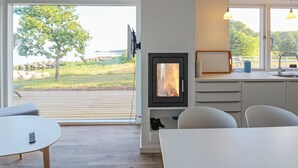 Een moderne keuken met een ingebouwde houtkachel, grote ramen met uitzicht op een boom en water, en een eethoek met een tafel en stoelen.