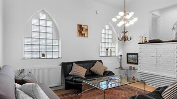 En moderne stue med en sort lædersofa, et glasbord og et tæppe med et mønster. Der er buede vinduer med persienner, en lysekrone og en pejs i hvide mursten.