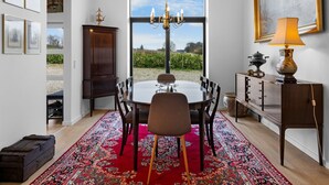 A dining area with a round table, chairs, a chandelier, a red carpet, and a view of vineyards through a large window.