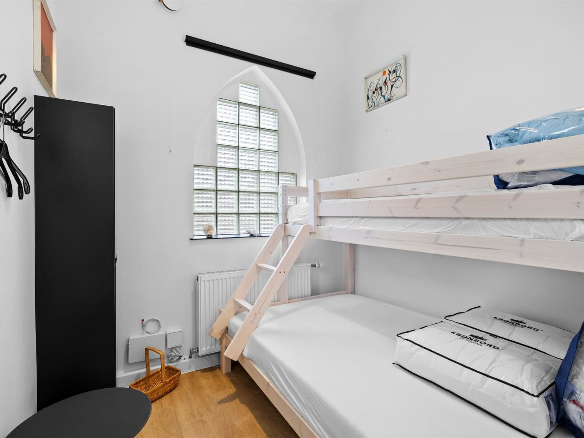 A bunk bed room with a window, a radiator, a black wardrobe, and a small table.