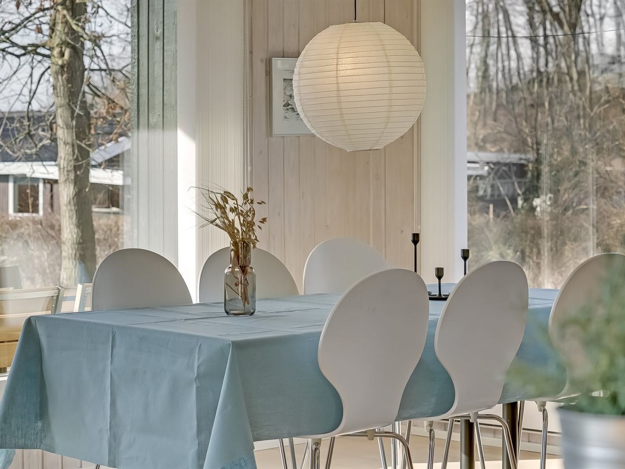 A dining table with a light blue tablecloth, surrounded by white chairs with curved backs. A clear glass vase with dried plants is placed on the table.